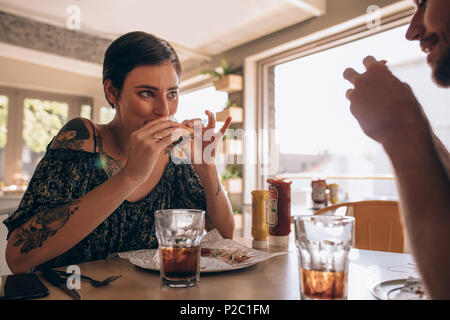 Young woman eating burger with her boyfriend sitting around the table at a restaurant. Couple having burger at cafe. Stock Photo