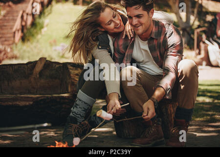 Happy man and woman sitting by fire and roasting marshmallows on sticks at campsite. Romantic couple having a great time on their camping. Stock Photo