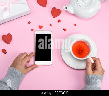left female hand holds a smartphone with a blank black screen, the left hand holds a cup with tea on a pink background, the top view Stock Photo