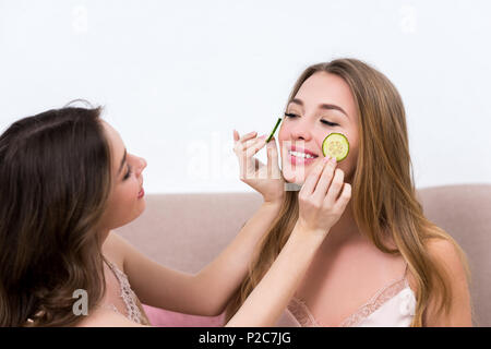 attractive girl holding slices of cucumber near face of smiling girlfriend Stock Photo