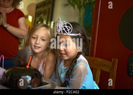Cute little preteen Asian girl wearing a princess dress and a tiara blowing out the candles on her birthday cake with her best friend sitting with her Stock Photo