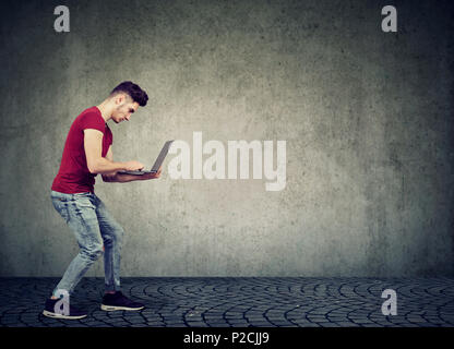 Side view of stylish young hipster man searching information in Internet using laptop against grunge wall. Stock Photo