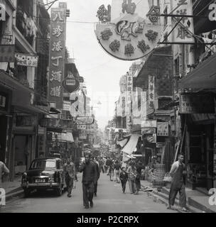 1950s, historical, a picture by J Allan Cash from this era looking down a street in the Old Town in Hong Kong, Asia, showing people walking in the street, stores, chinese signs and the architecture of the area, with the living accomodation above the retail outlets. Also in the picture, parked in the street, a motorcar of the era with a number plate, 423. Stock Photo