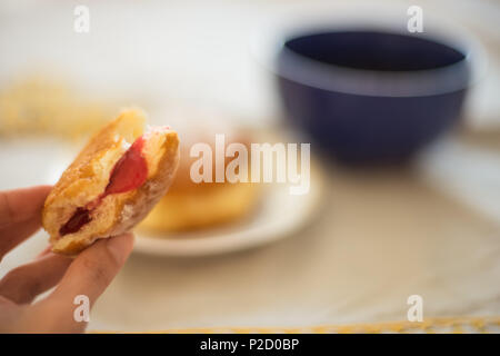bitten donut with a pink jam in hand on the background of a table with a mug of tea Stock Photo