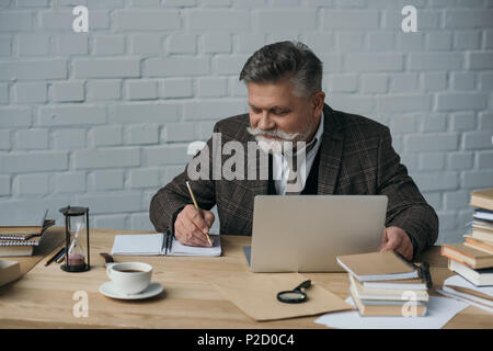 happy senior writer working with laptop and making notes in notebook Stock Photo