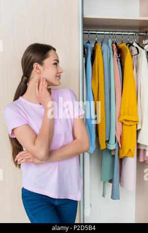 thoughtful young woman looking at clothes hanging in cabinet at home Stock Photo