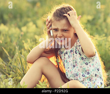 Angry serious cute kid girl sitting on the glass, talking on mobile phone and scratching the head on summer green grass background. Toned closeup fun  Stock Photo