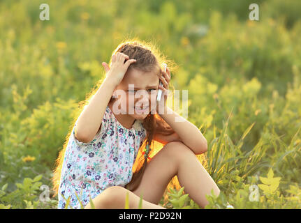 Angry serious cute kid girl sitting on the glass, talking on mobile phone and scratching the head on summer green grass background. Stock Photo