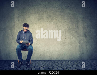 Stylish young man sitting on chair and browsing tablet with confidence on gray wall background Stock Photo