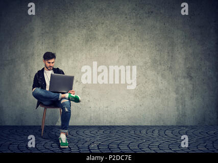 Stylish young man sitting on chair and browsing laptop with confident on gray simple background Stock Photo