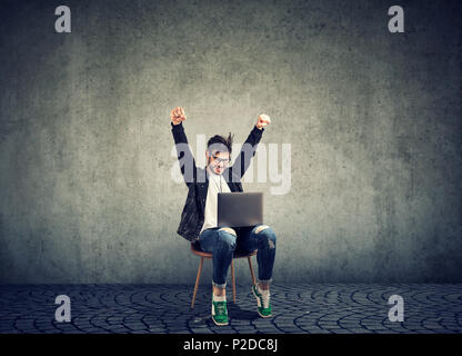 Stylish hipster man sitting on chair with laptop and celebrating victory looking excited with new achievement on gray Stock Photo