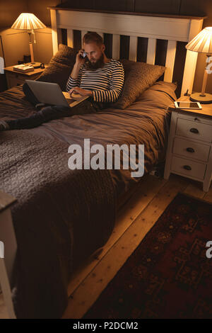 Man lying on bed talking on phone while using laptop Stock Photo