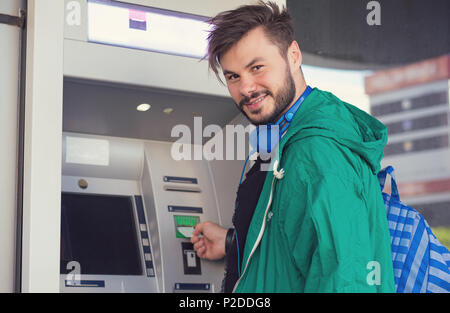 Young hipster guy in headphones smiling at camera while inserting credit card in ATM machine getting cash money Stock Photo
