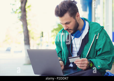 Stylish hipster man sitting on street with laptop on knees and doing online purchase with credit card Stock Photo