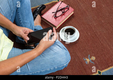 Close up of young woman wearing casual clothing and necklace sitting on carpet,  holding a  smartphone and red vintage book, a cup of coffee, and blac Stock Photo