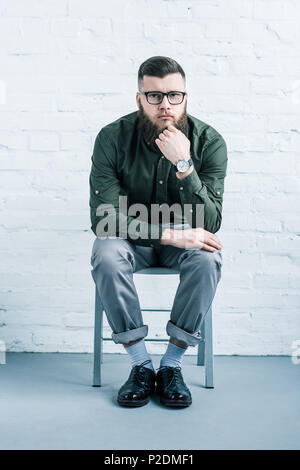 thoughtful stylish businessman sitting on chair against white brick wall Stock Photo