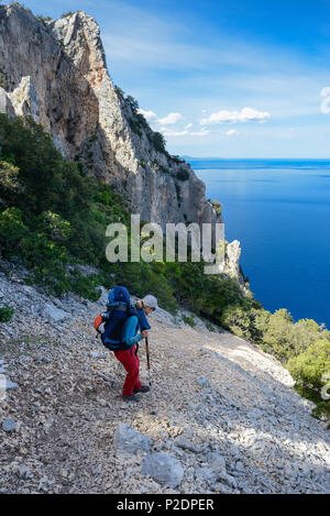 A young woman with trekking gear hiking down a steep slope along the mountainous coast above the sea, Golfo di Orosei, Selvaggio Stock Photo