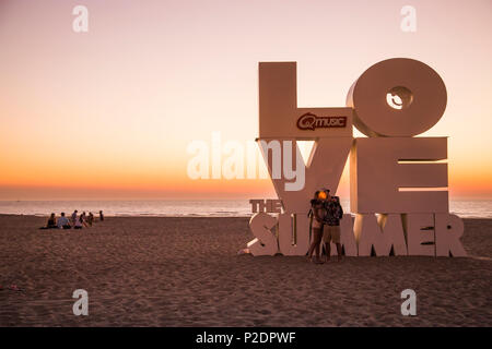 Couple taking a selfie photograph with smartphone in front of Love sculpture on the beach at sunset, Ostend, Flanders, Flemish R Stock Photo