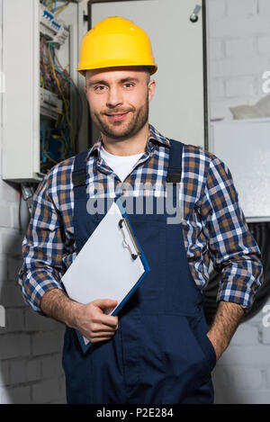 Electrician holding clipboard and smiling by electrical box Stock Photo