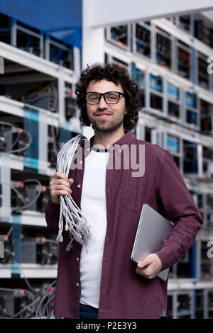 handsome young computer engineer with laptop and wires working at cryptocurrency mining farm Stock Photo