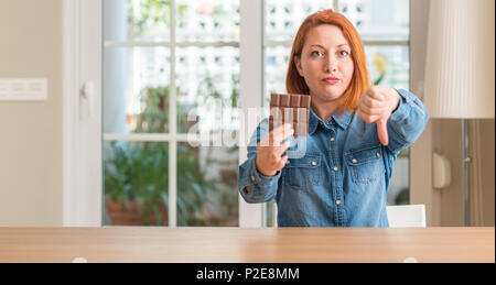 Redhead woman holding chocolate bar at home with angry face, negative sign showing dislike with thumbs down, rejection concept Stock Photo