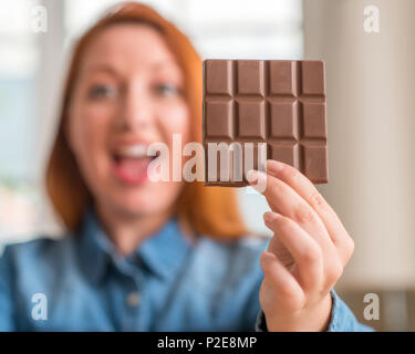 Redhead woman holding chocolate bar at home very happy and excited, winner expression celebrating victory screaming with big smile and raised hands Stock Photo