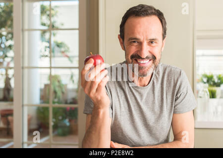 Middle age man eating healthy red apple with a happy face standing and smiling with a confident smile showing teeth Stock Photo