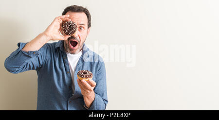 Senior man eating chocolate donut scared in shock with a surprise face, afraid and excited with fear expression Stock Photo