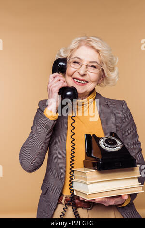 happy fashionable senior woman in eyeglasses talking on rotary phone and holding stack of books Stock Photo
