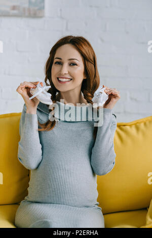 smiling pregnant woman holding newborn shoes and looking at camera in living room Stock Photo