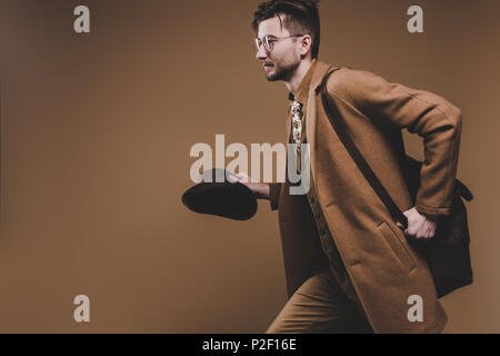 Young man in coat holding hat and running isolated on brown Stock Photo