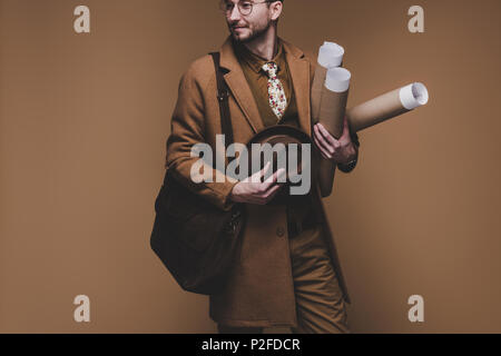 Young man in vintage styled clothes holding rolled papers isolated on brown Stock Photo