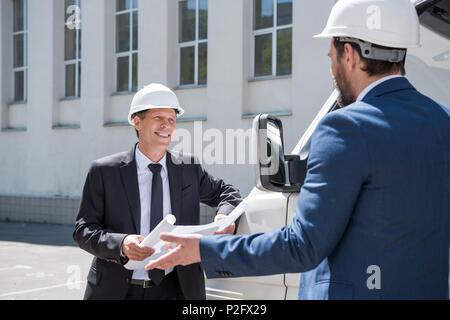 Smiling architect in hardhat holding blueprint and looking at colleague Stock Photo