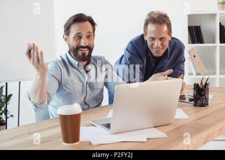 Two smiling businessmen using laptop and drinking coffee from paper cup in office Stock Photo