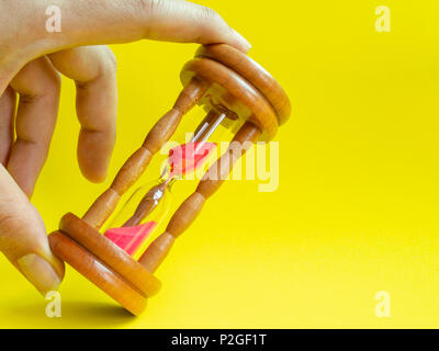 Hands Holding The Wooden Vintage Hourglass with Pink Sand on Yellow Background, Sand Running through the Bulbs for Measuring The Passing Time in A Cou Stock Photo