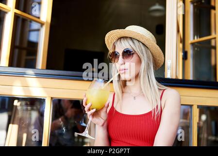 Young pretty sympathetic blonde sitting in a cafe near an open window with a cocktail in her arms and in a straw hat. Stock Photo