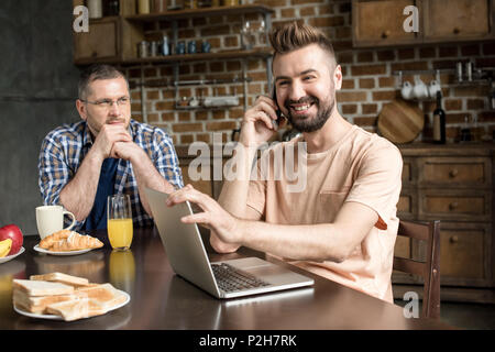 Smiling bearded man using laptop and talking on smartphone while pensive partner sitting at table behind Stock Photo