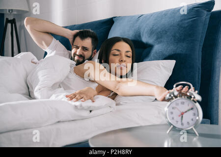 Young couple trying to turn off alarm clock while sleeping in bed Stock Photo