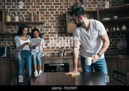 father drinking coffee and cleaning table while mother and daughter using digital tablet at kitchen Stock Photo