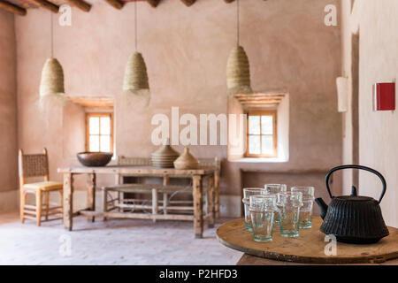 Cast iron tea pot and glasses in foreground of moroccan dining room with rattan oendant lights, earth walls and terracotta floors Stock Photo