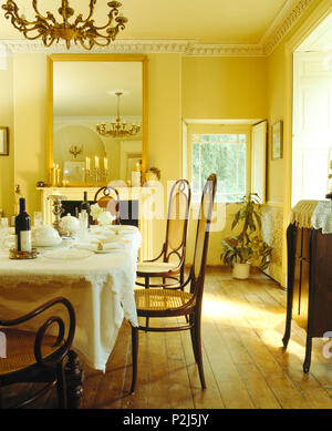 Antique Bentwood chairs at table set for lunch in yellow dining room with large mirror above fireplace and wooden flooring Stock Photo