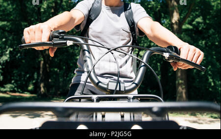 closeup of a young caucasian man, carrying a backpack, seen from the front, riding a bike by a public park Stock Photo