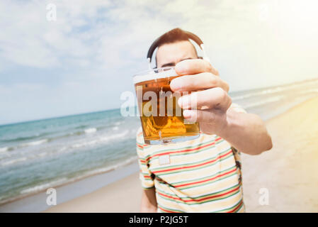 closeup of a young caucasian man on the beach listening to music with a pair of wireless headphones while is drinking a beer in front of the sea, with Stock Photo