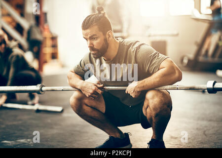 Fit young man in sportswear looking focused while crouching on a gym floor holding a barbell during a workout session Stock Photo