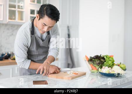 Asian confused man reading his tablet with a pensive thoughtful look while standing in his kitchen while cooking and preparing a meal from a variety o Stock Photo