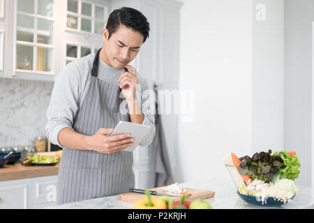 Asian confused man reading his tablet with a pensive thoughtful look while standing in his kitchen while cooking and preparing a meal from a variety o Stock Photo