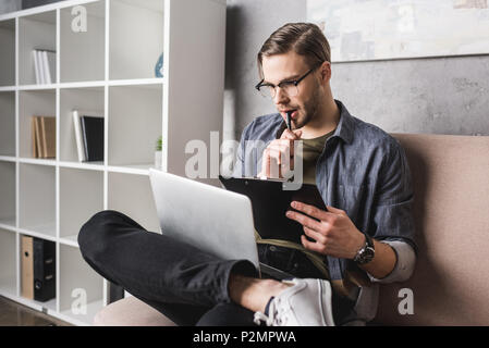 young thoughtful man working with laptop on couch and making notes on clipboard Stock Photo