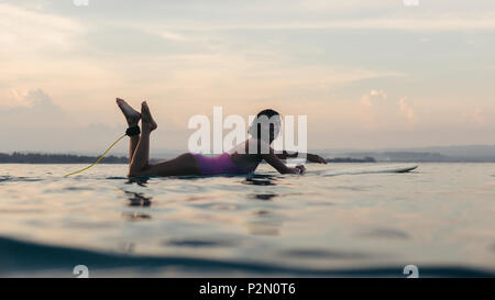 silhouette of woman lying on surfboard in water in ocean at sunset Stock Photo