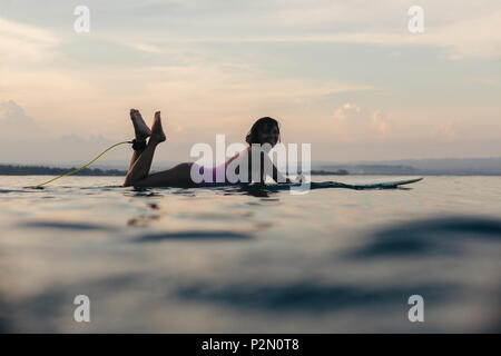 silhouette of smiling girl lying on surfboard in ocean at sunset Stock Photo