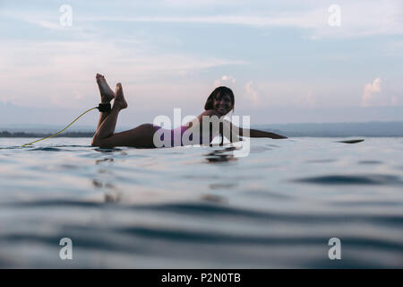 smiling woman lying on surfboard in water in ocean at sunset Stock Photo